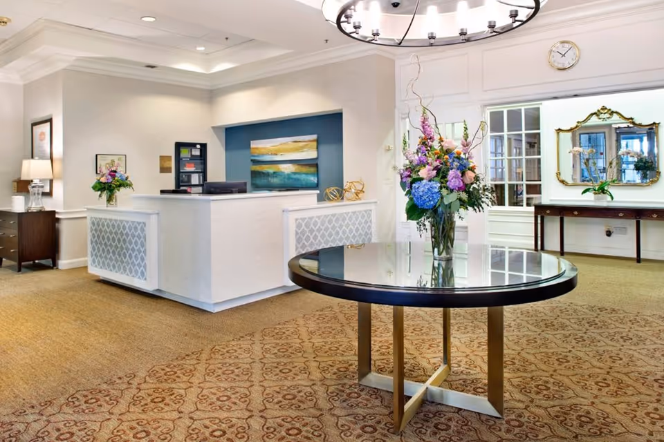 Bright reception lobby with a round glass-top table holding a large floral arrangement and a check-in desk in the background.