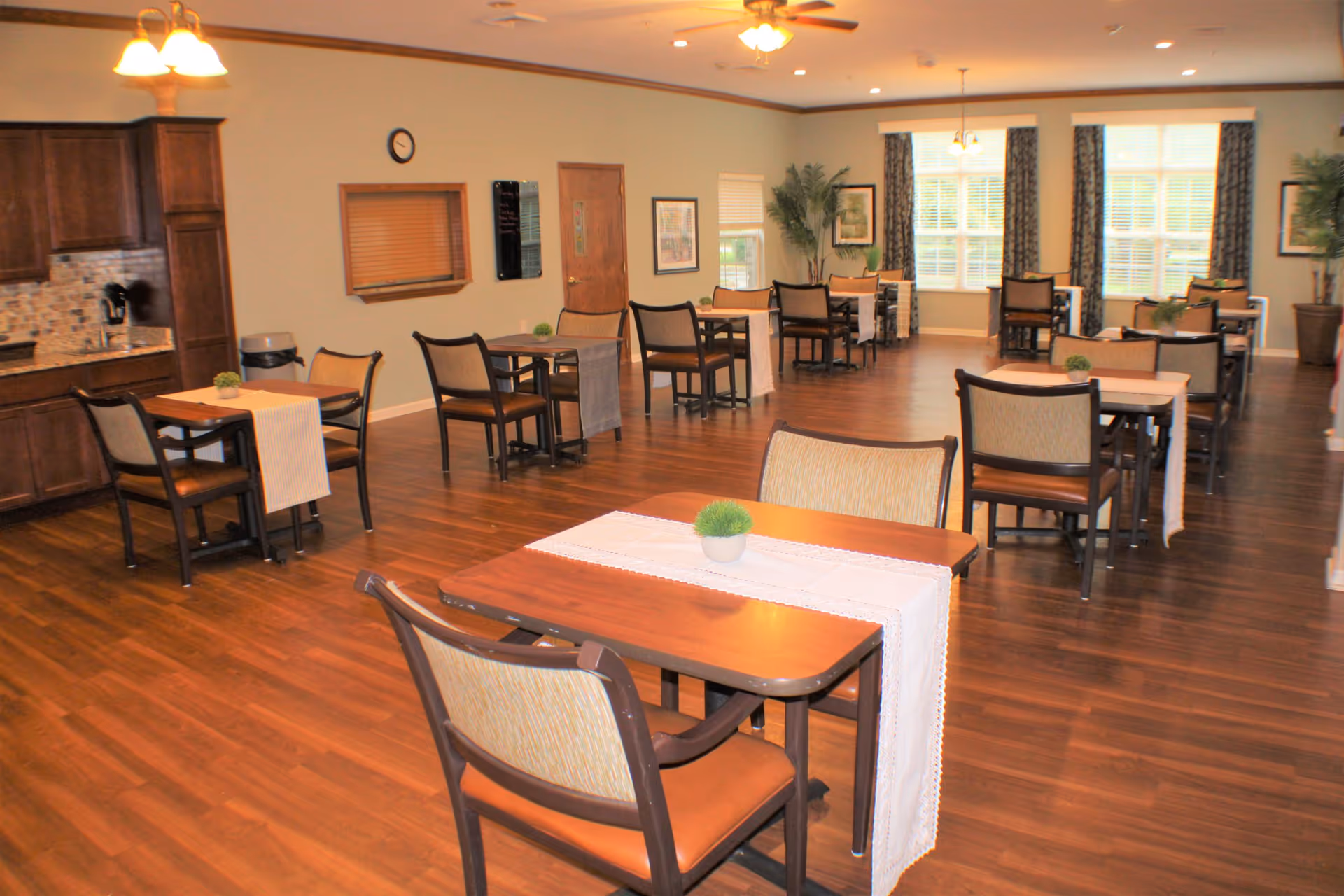 A spacious dining room with multiple wooden tables and chairs arranged neatly. Each table has a white table runner and a small green plant centerpiece. The room features wood flooring, large windows with curtains allowing natural light, and a ceiling fan with lights. There is a kitchen area with wooden cabinets and a tiled backsplash on the left side of the room.