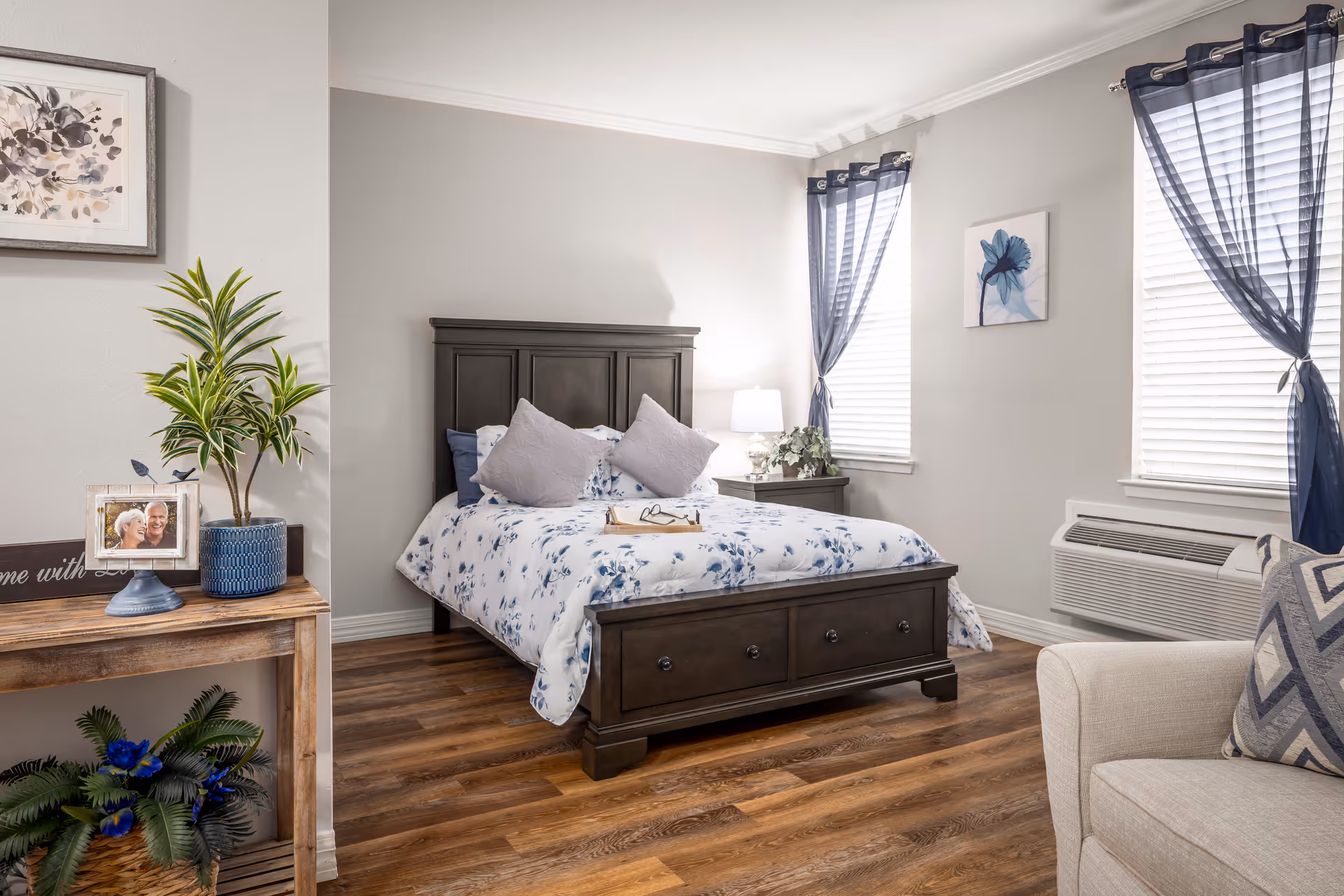 A cozy bedroom with a dark wooden bed frame and floral bedding, two light gray pillows, a nightstand with a lamp and plant, two windows with blue sheer curtains, a beige armchair with a patterned pillow, and a wooden console table with a potted plant, framed photo, and decorative sign.