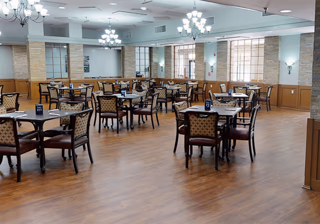 Spacious dining room with multiple tables and patterned chairs arranged on wood flooring beneath chandeliers.