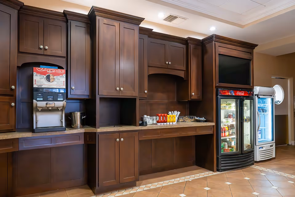 Interior view of a beverage and snack station with dark wooden cabinets, a coffee machine, condiment containers, and two glass-door refrigerators stocked with drinks and snacks in a senior living facility.
