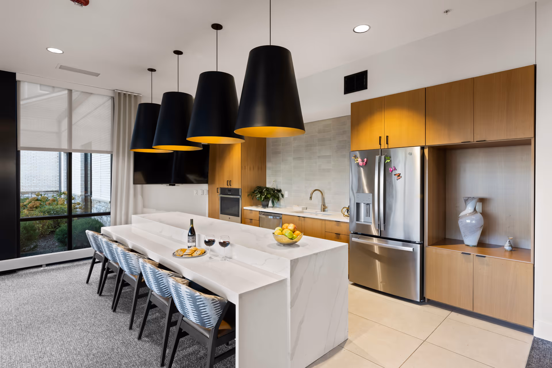 Modern kitchen area with a long white marble island featuring six chairs, three large black pendant lights hanging above, wooden cabinets, a stainless steel refrigerator, built-in oven, and a window showing an outdoor garden area.