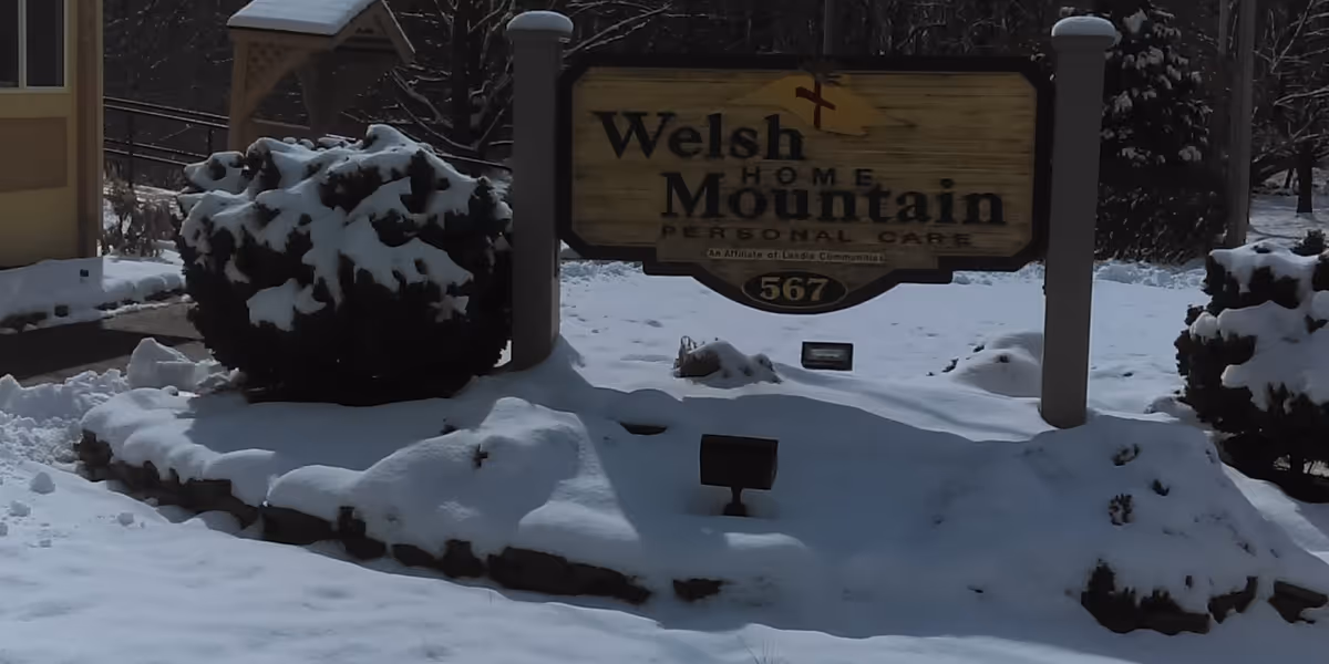 A wooden sign for Welsh Mountain Home Personal Care covered with snow, surrounded by snow-covered bushes and ground, indicating the facility's location at 567.