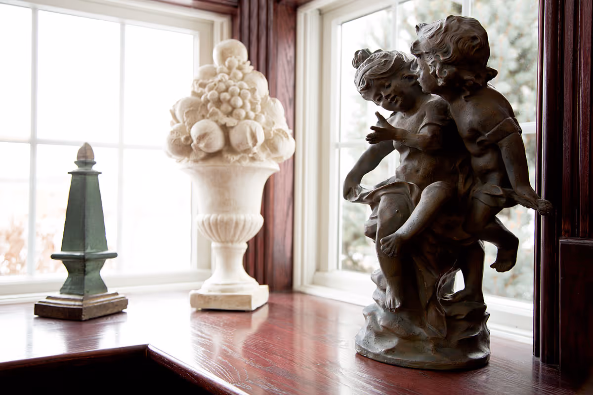 A wooden windowsill with three decorative sculptures: a green obelisk, a white vase filled with fruit, and a bronze statue of two children playing, set against a window with white frames.