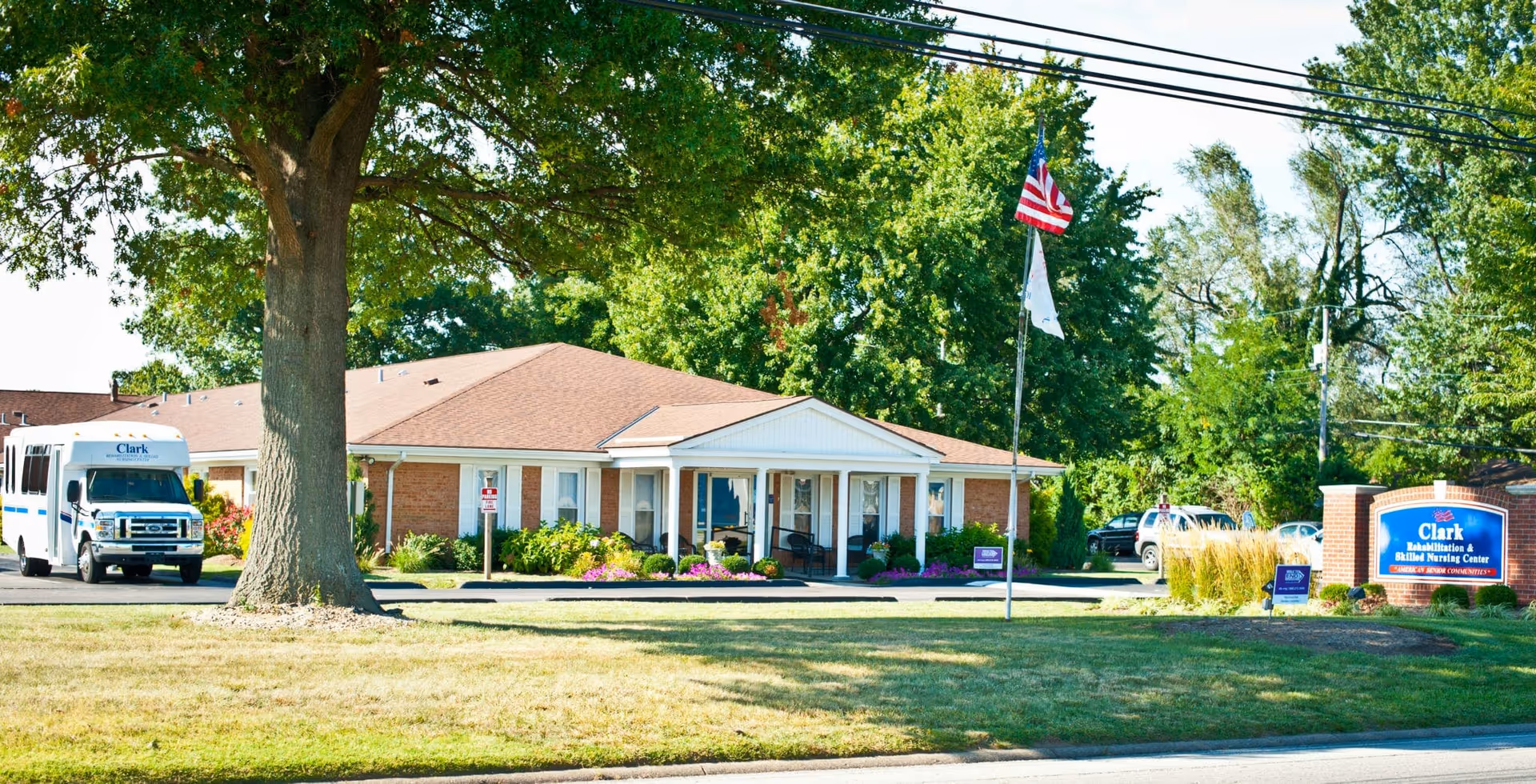 Exterior view of Clark Rehabilitation and Skilled Nursing Center building with a large tree in the foreground, an American flag on a flagpole, a shuttle bus parked on the left side, and a blue facility sign on the right side surrounded by greenery and flowers.