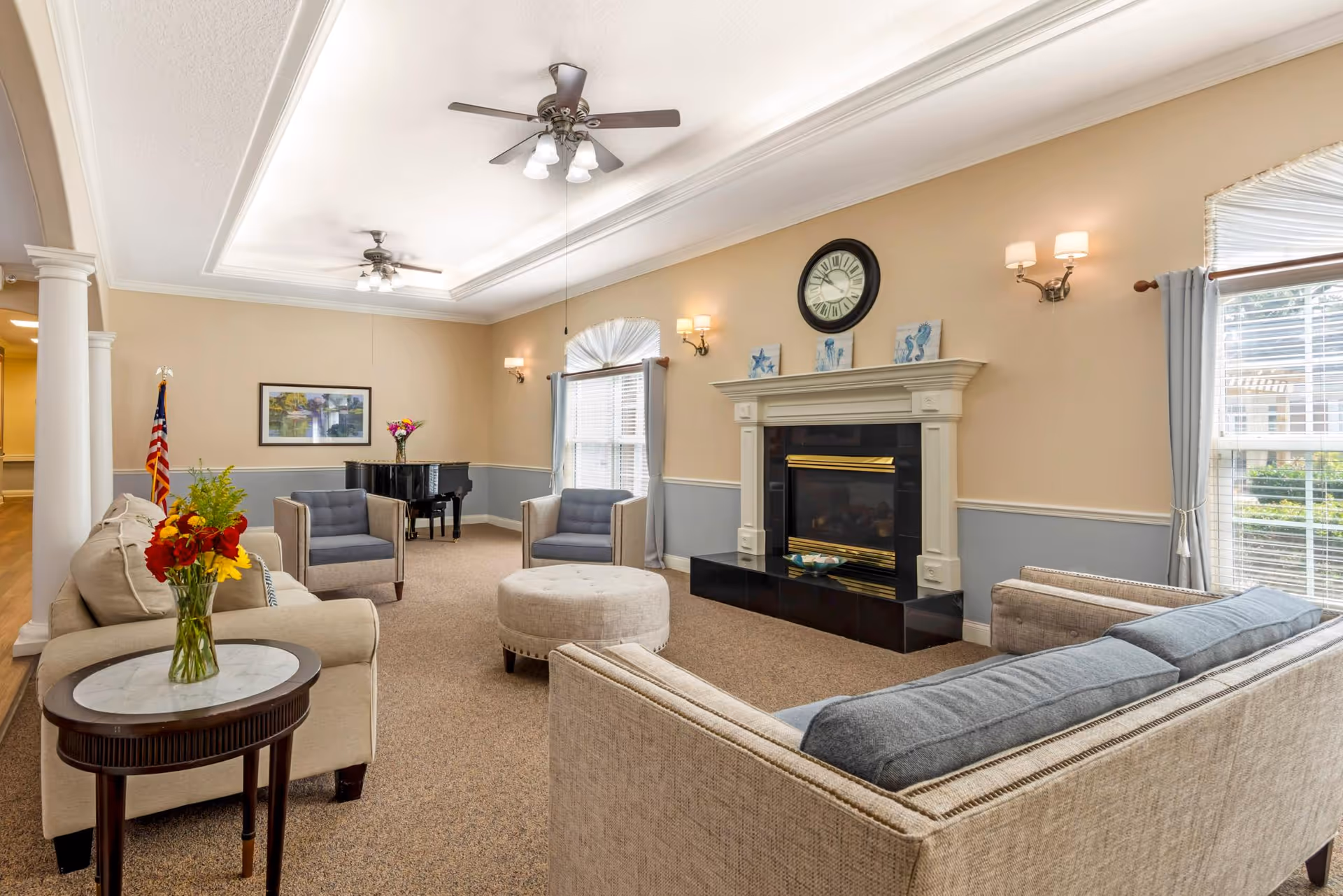 A spacious living room with beige walls and carpeted floor, featuring a fireplace with a clock above it, two armchairs, a beige sofa, a round ottoman, and a side table with a vase of flowers. There is a grand piano in the corner near a window with curtains, and ceiling fans with lights hang from the tray ceiling.