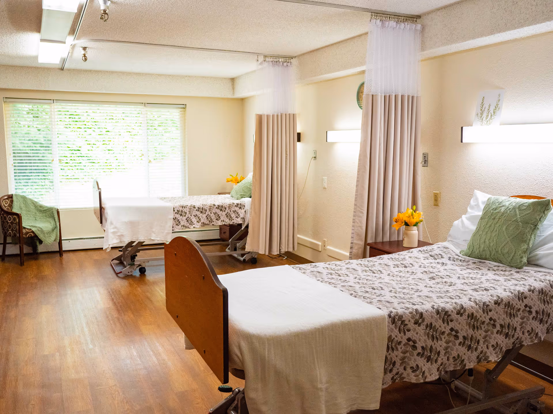 A bright room with two hospital-style beds separated by privacy curtains. Each bed is made with floral-patterned bedding and a green pillow. There is a wooden chair with a green blanket near a large window with blinds. The room has wooden flooring and soft lighting from wall-mounted fixtures.