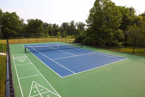 Outdoor tennis court with a blue playing surface and green surrounding area, enclosed by a black chain-link fence, with trees and grass in the background under a clear sky.