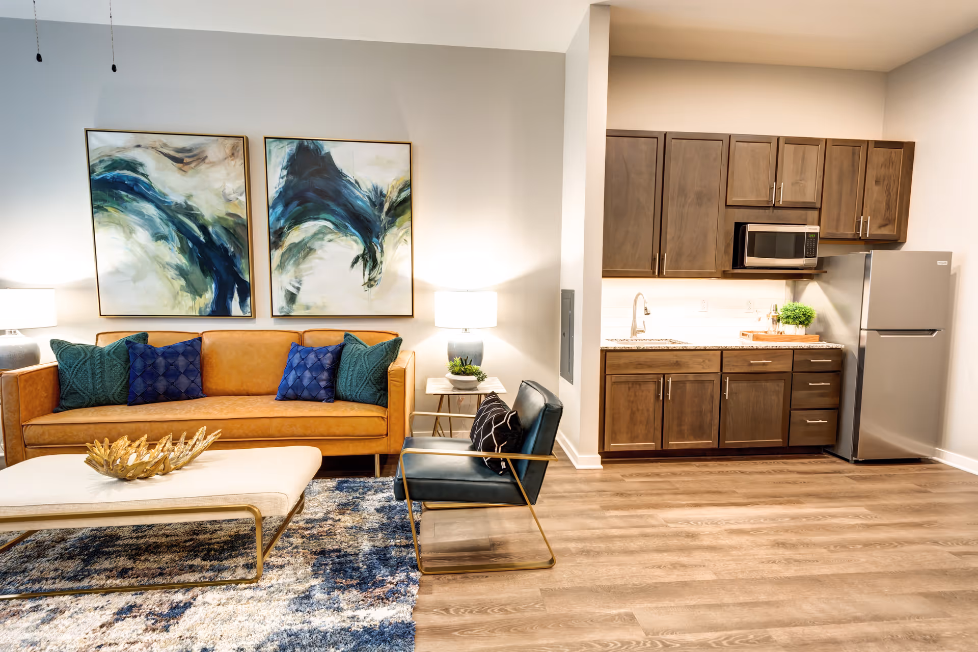 A modern senior living space featuring a tan leather sofa with blue and green pillows, two abstract paintings on the wall above the sofa, a blue armchair with a black and white pillow, a beige ottoman with a decorative gold centerpiece, and a small kitchen area with wooden cabinets, a stainless steel refrigerator, microwave, and sink. The floor is wood, and the room is well-lit with table lamps.