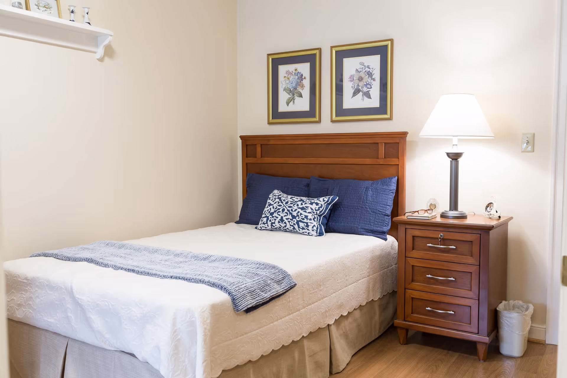 A neatly made bed with a wooden headboard, white bedspread, blue pillows, and a blue throw blanket. Next to the bed is a wooden nightstand with three drawers, a lamp, a pair of glasses, and a small clock. Two framed floral pictures hang on the wall above the headboard. The room has light-colored walls and wooden flooring.