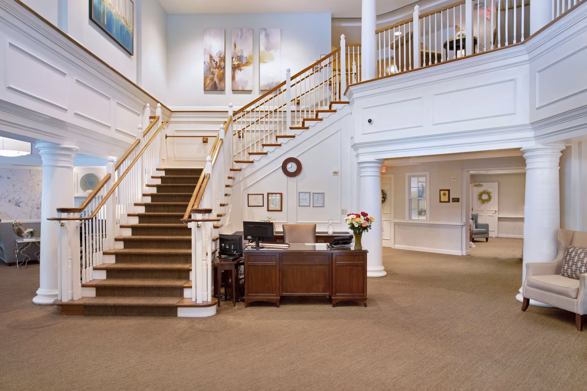 Interior view of a senior living facility lobby with a central staircase featuring white railings and wooden handrails. A wooden reception desk with a computer and a vase of flowers is positioned in front of the staircase. The area is carpeted and decorated with framed artwork and certificates on the walls. There are white columns and a seating area with chairs visible in the background.