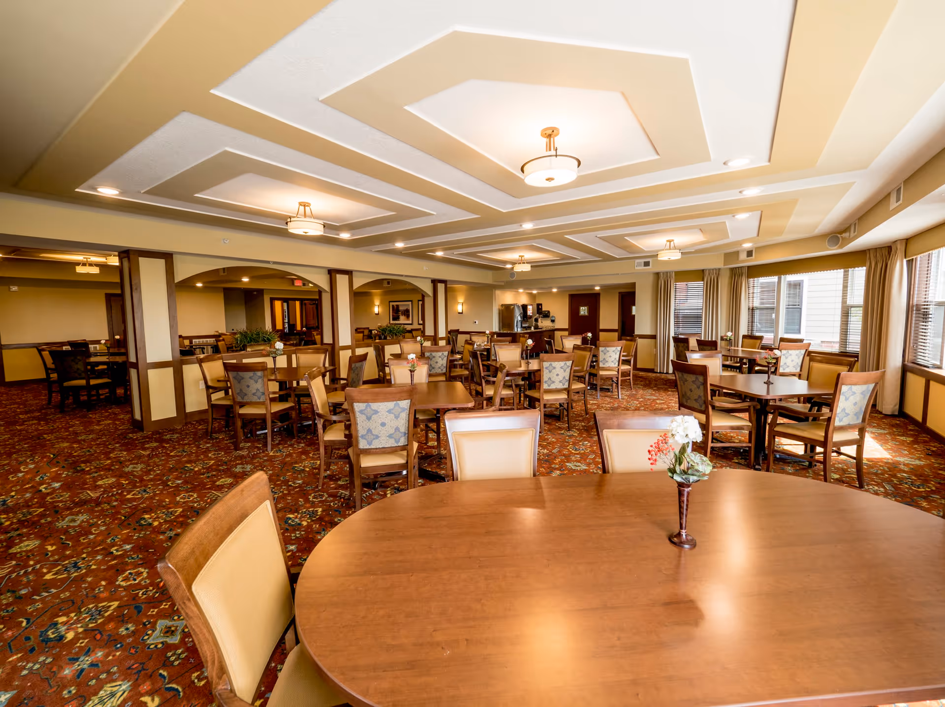 Spacious senior living dining room with multiple wooden tables and chairs, decorative ceiling lights, and floral centerpieces.