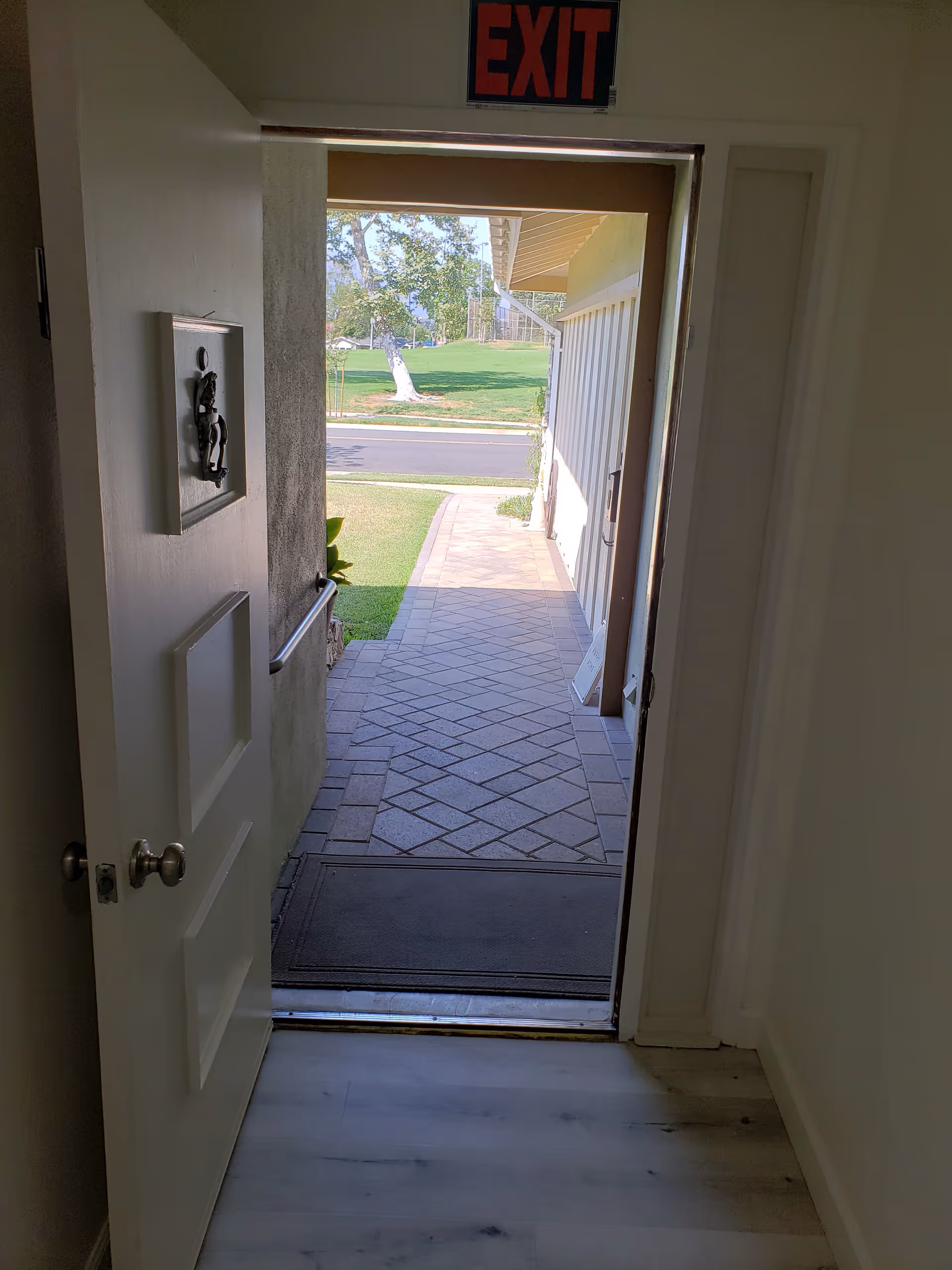 View through an open door leading outside to a paved walkway and green lawn with trees, with an illuminated red EXIT sign above the door.
