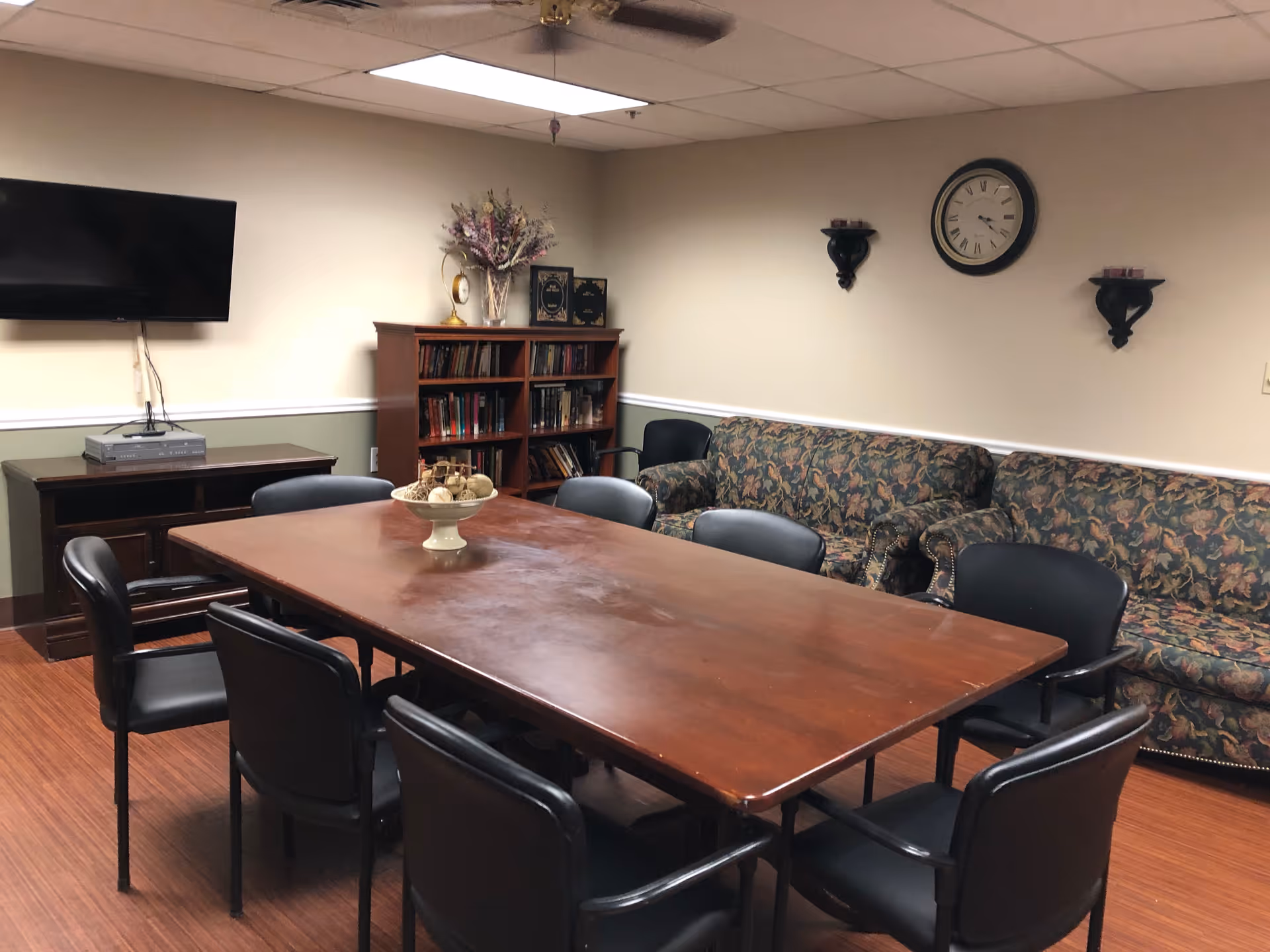 A meeting or common room with a large wooden table surrounded by eight black chairs. In the background, there are two floral-patterned couches, a wooden bookshelf filled with books, a wall clock, and two wall-mounted candle holders. A flat-screen TV is mounted on the wall above a wooden cabinet. The room has a ceiling fan and wood flooring.