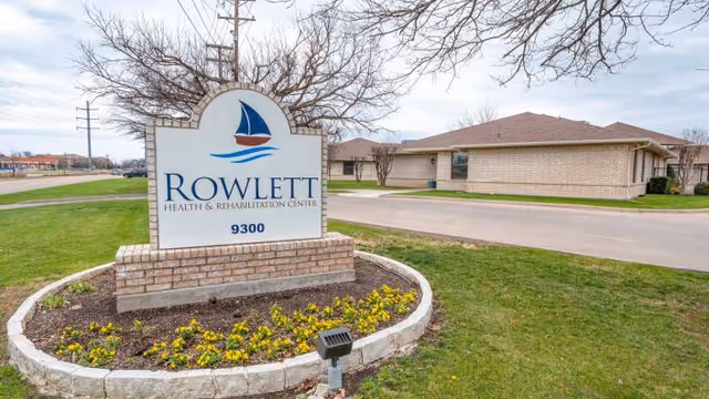 Outdoor view of Rowlett Health & Rehabilitation Center sign with a sailboat logo, located on a grassy area with yellow flowers planted around the base. The building and driveway are visible in the background under a cloudy sky.