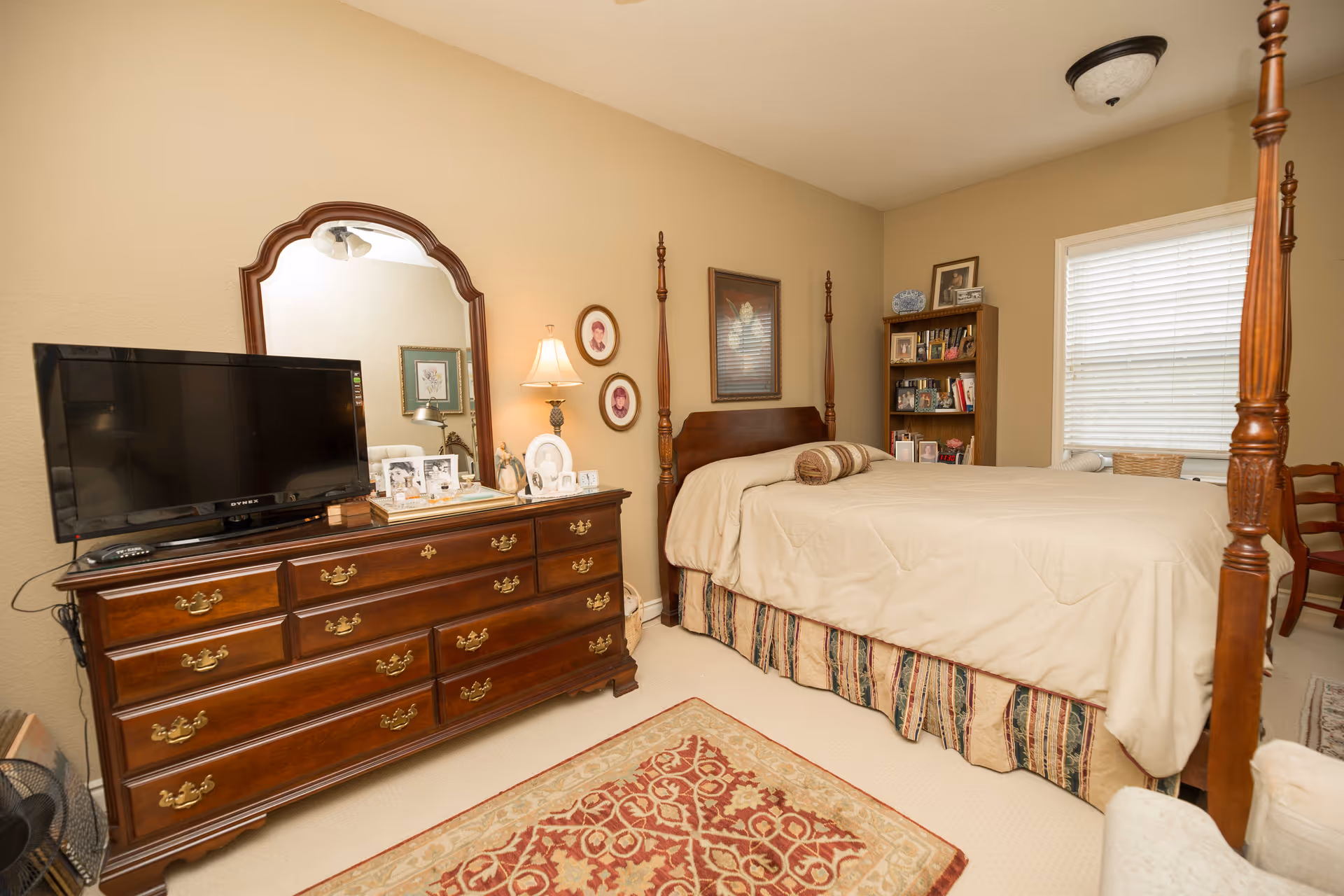 Well-appointed bedroom with a four-poster bed, wooden dresser with mirror and TV, bookshelf, and a window with blinds.