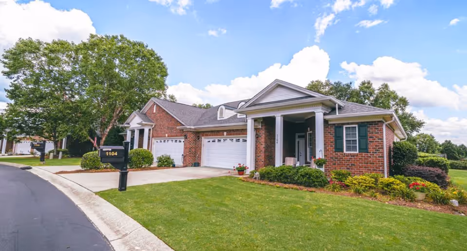 A single-story brick house with a white garage door and a covered front porch with white columns. The house is surrounded by a well-maintained lawn, shrubs, and flowers. A black mailbox with the number 1104 is visible near the driveway. The sky is partly cloudy with blue patches.