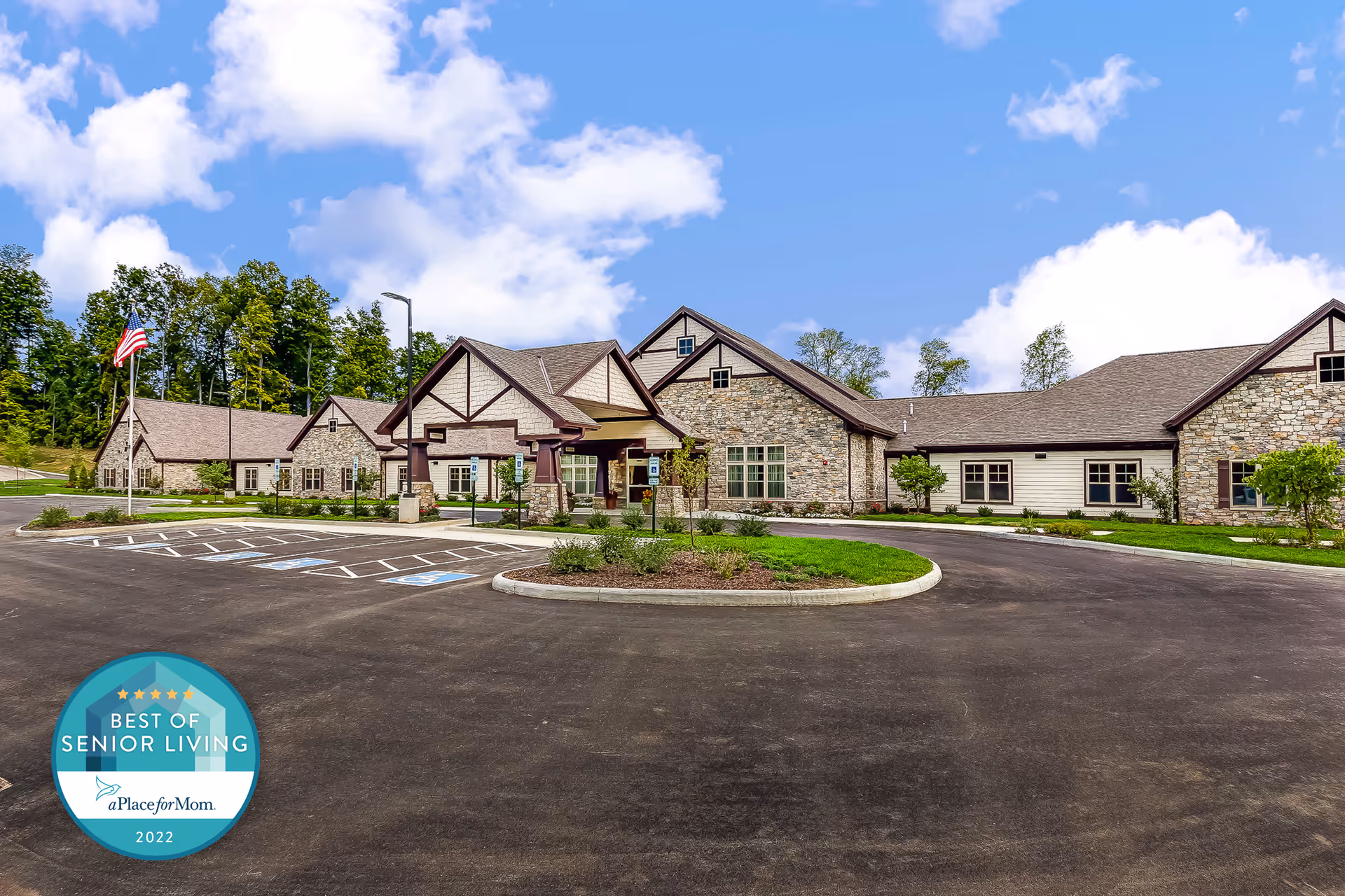 Exterior view of a senior living facility building with stone and wood siding, a covered entrance, and a circular driveway with parking spaces including handicapped spots. There is an American flag on a flagpole to the left and trees in the background under a partly cloudy blue sky.