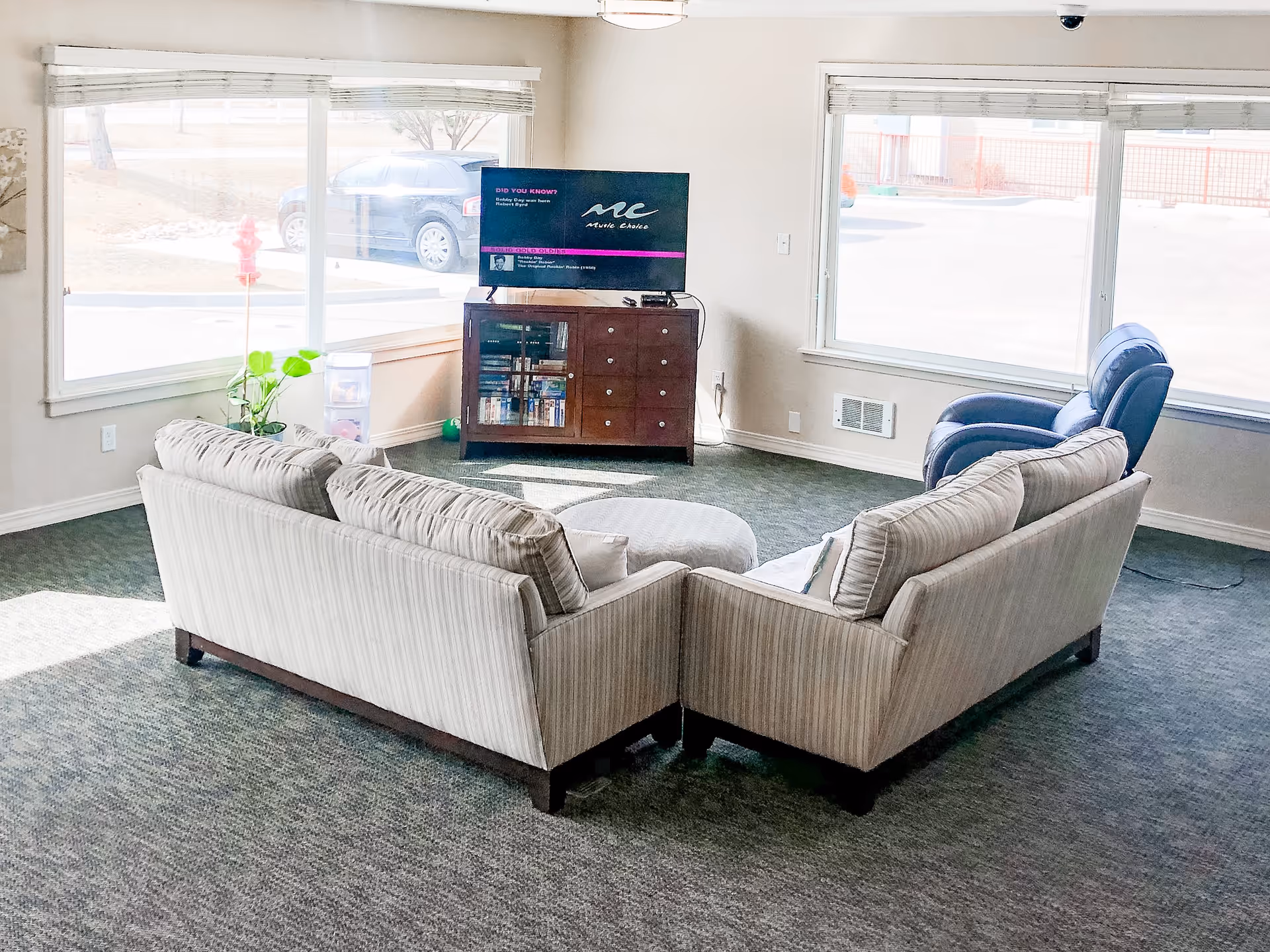 Communal living room with two beige sofas and an ottoman arranged facing a television on a wooden cabinet beneath large windows.