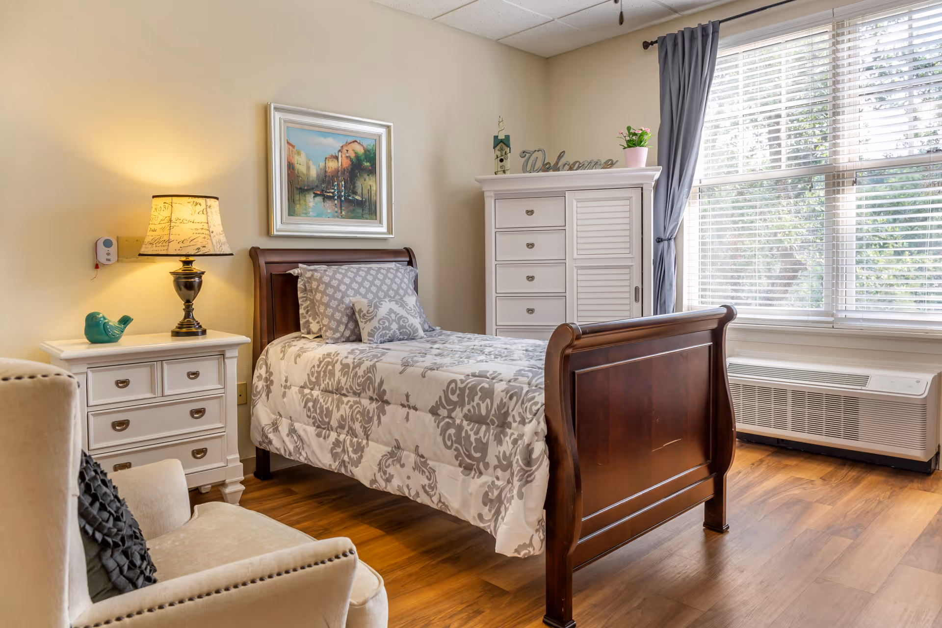 A cozy bedroom with a single wooden bed featuring a patterned gray and white comforter and pillows. Next to the bed is a white nightstand with a decorative lamp and a small blue bird figurine. A white dresser with drawers and a cabinet stands against the wall, topped with a 'Welcome' sign and a small potted plant. A large window with white blinds and gray curtains allows natural light to fill the room. A beige armchair with a black cushion is partially visible in the foreground.