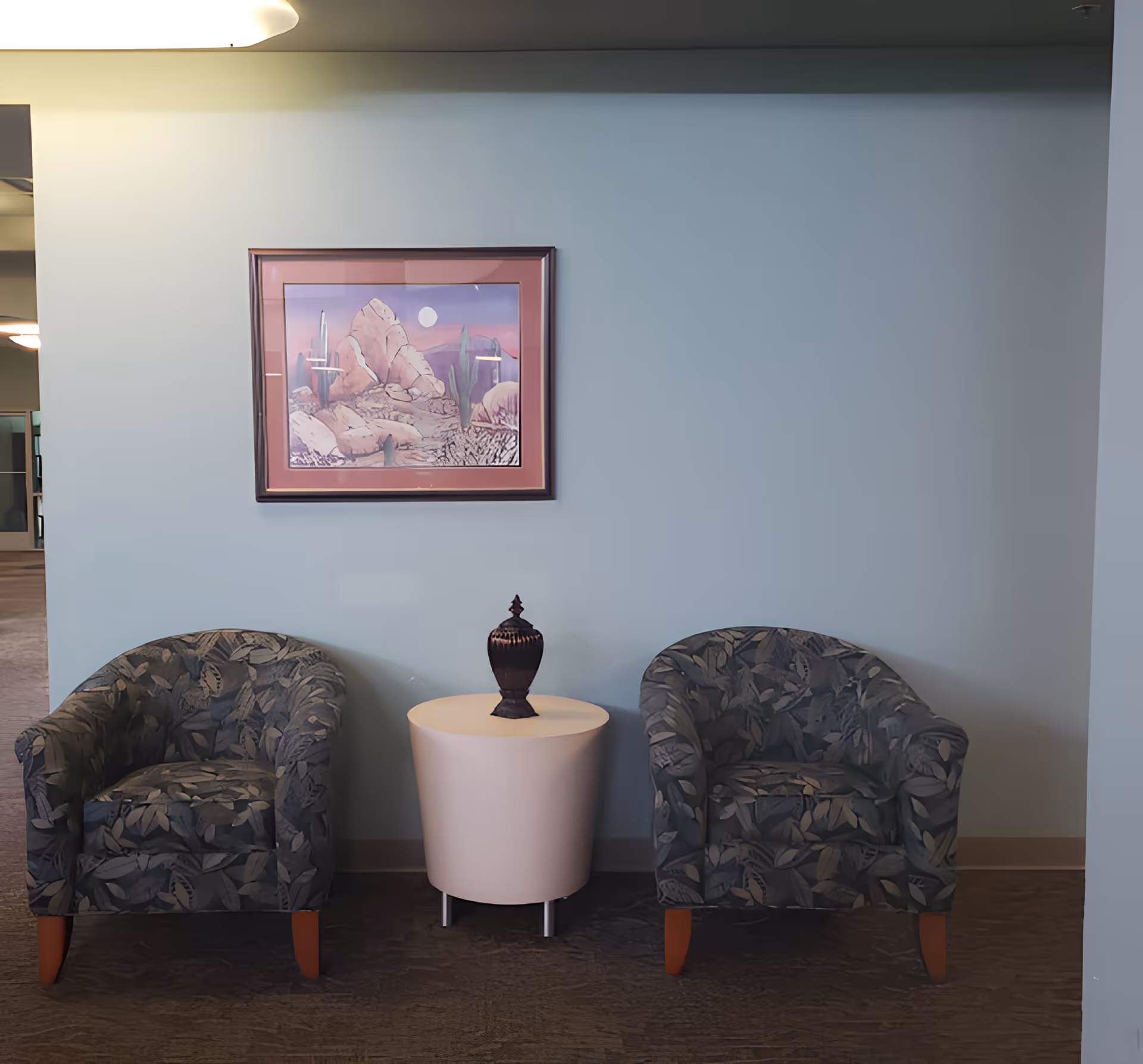 Two patterned armchairs with wooden legs are placed on either side of a small round white table with a decorative vase on top. Above the table, a framed picture of a desert landscape with rocks and cacti is hung on a light blue wall. The setting appears to be an indoor waiting or lounge area.