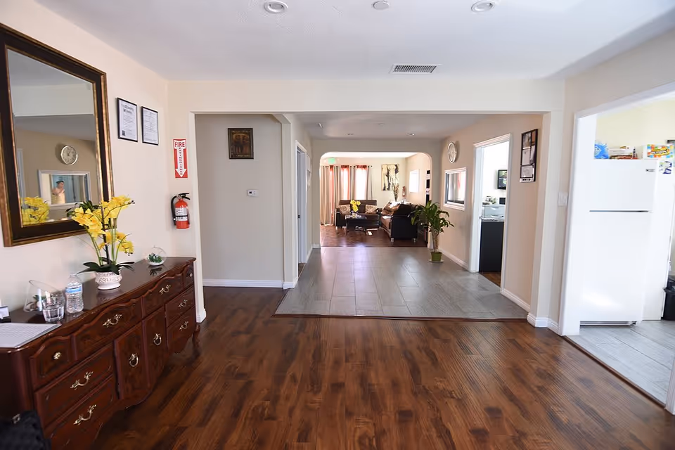 Interior view of a senior care facility showing a hallway with wooden flooring leading to a living room area with sofas and a coffee table. To the right, there is a kitchen area with a white refrigerator. On the left side of the hallway, there is a wooden dresser with yellow flowers in a vase and a large mirror above it. The walls are light-colored, and there is a fire extinguisher mounted on the wall.