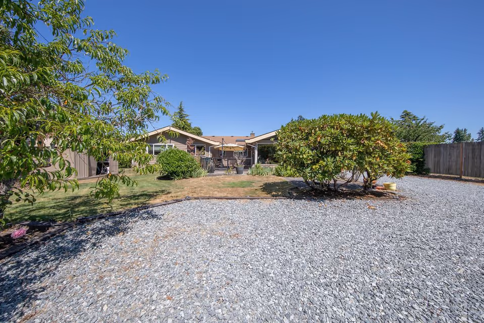 Outdoor view of a single-story residential building with a gravel driveway in the foreground, green bushes and trees on either side, and a clear blue sky above.