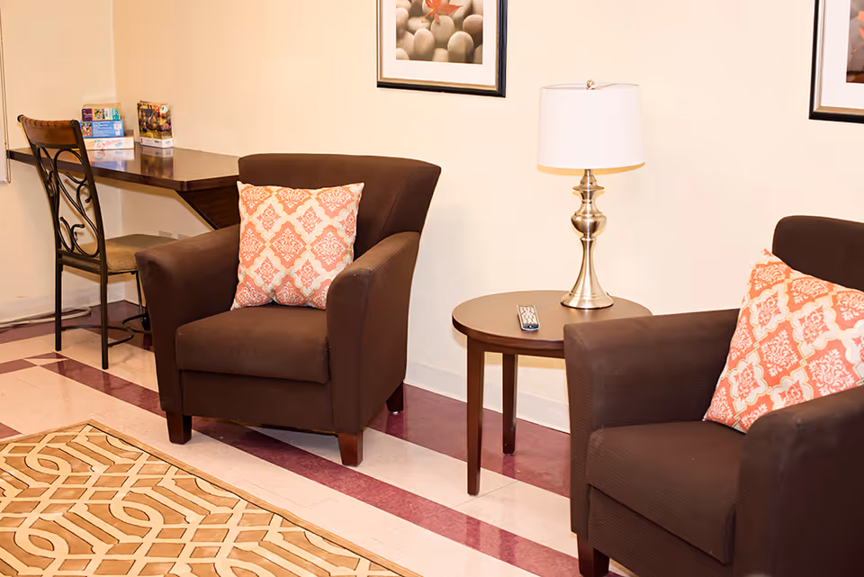 Two brown armchairs with patterned cushions flank a small round table topped by a lamp in a sitting area with a desk and chair in the background.