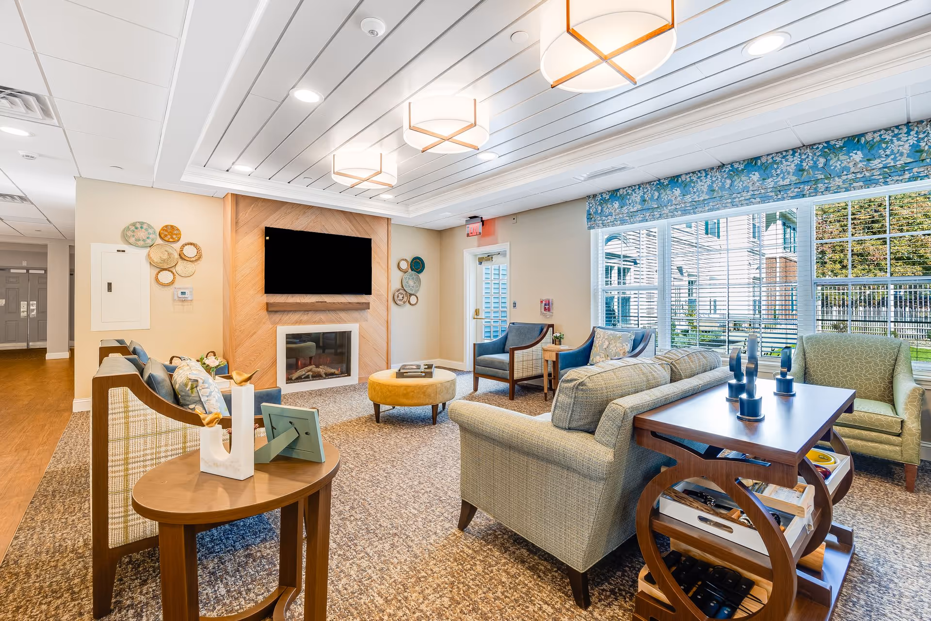 Bright, well-appointed common living room with sofas and chairs arranged around a fireplace and wall-mounted TV, large windows overlooking a courtyard, and modern ceiling lights.