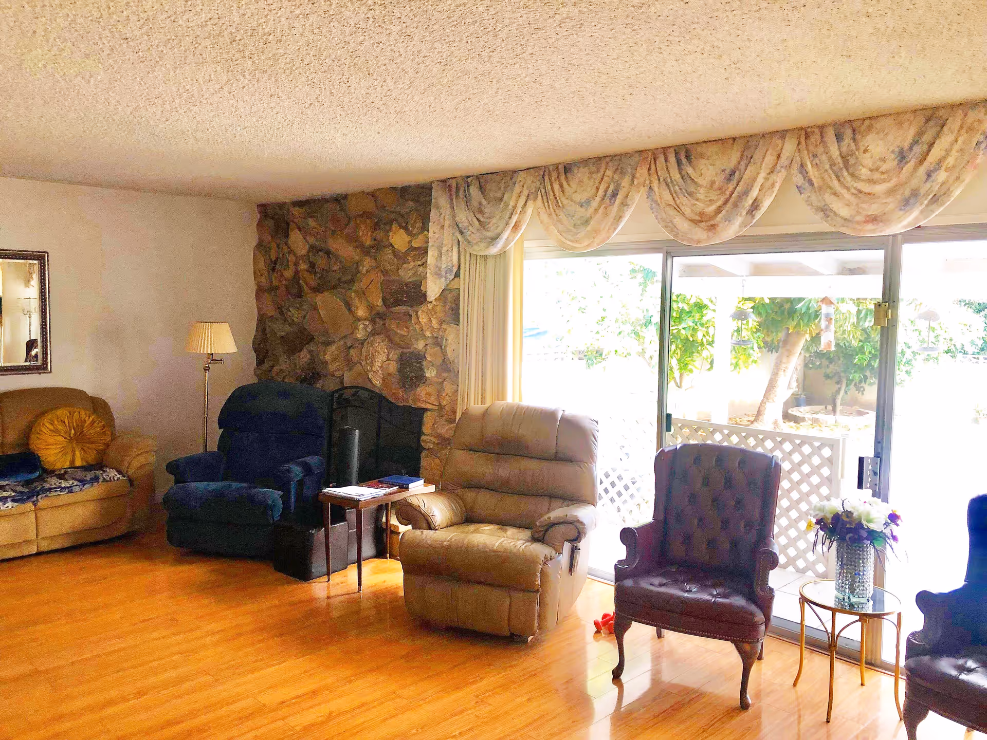 Sunlit living room with several armchairs and recliners on hardwood floors in front of sliding glass doors and a stone fireplace.