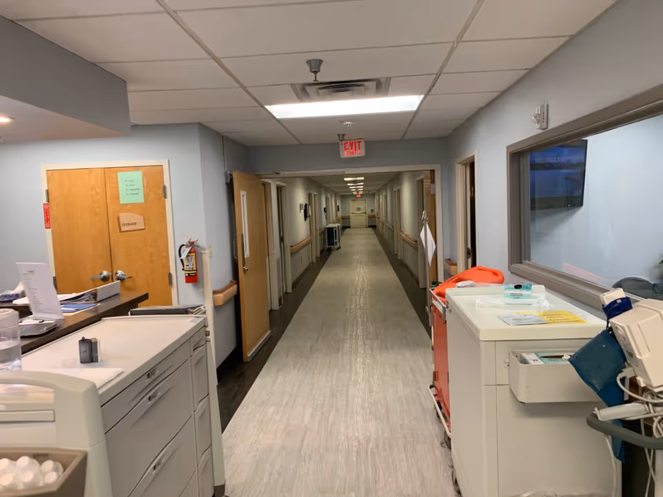 A long, clean hallway in a healthcare facility with light gray walls and a light-colored floor. On the left side, there is a counter with papers and a water bottle, and a wooden door labeled 'Storage'. On the right side, there is medical equipment and a window looking into another room with a television screen. The hallway has handrails on both sides and an exit sign at the far end.