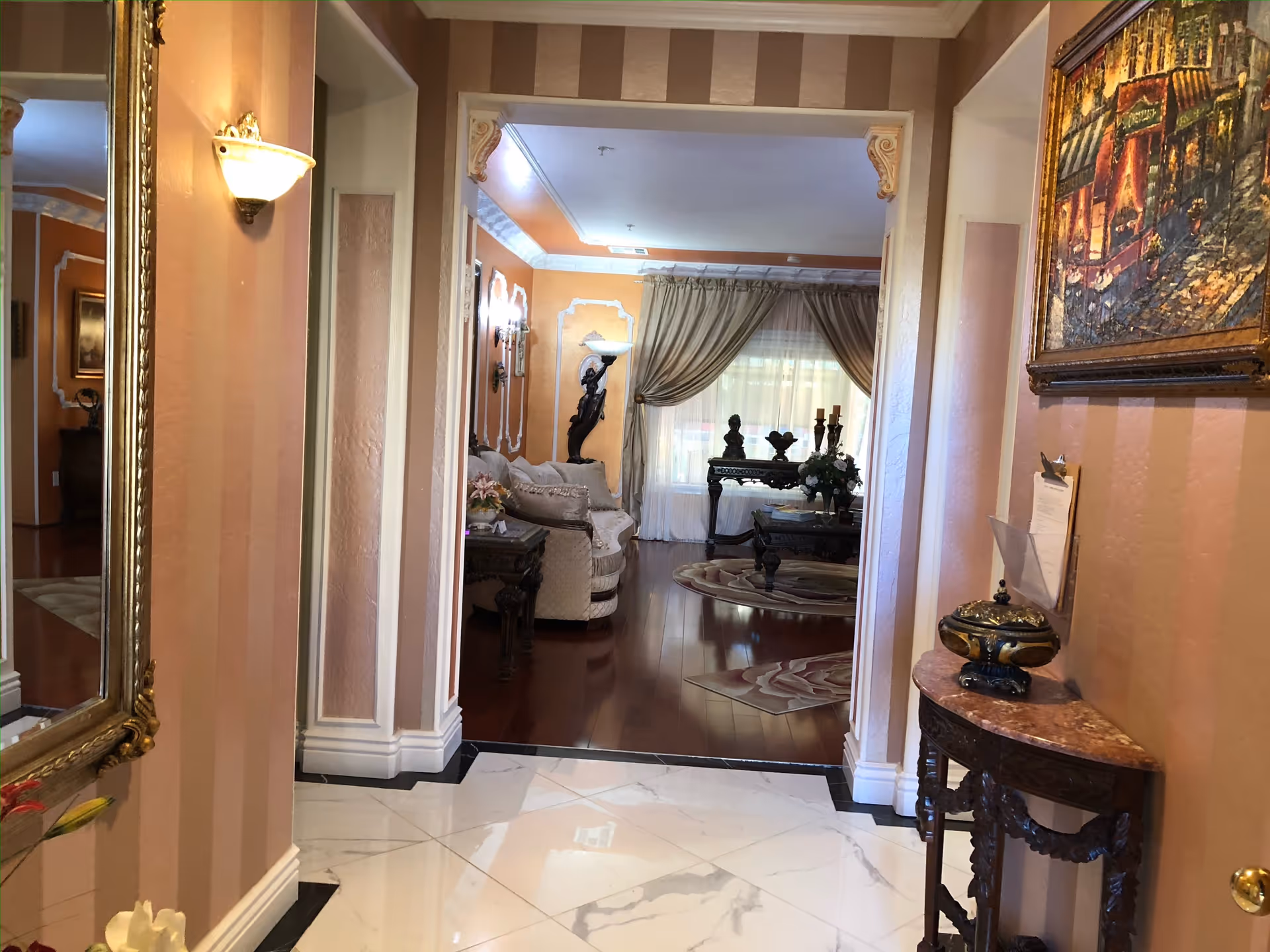 View of an elegant interior hallway leading into a living room area. The hallway has polished white marble flooring with black accents, peach-colored striped walls, and a large ornate mirror on the left wall. The living room features wooden flooring, a beige sofa, a decorative table with sculptures and flowers, and large windows with beige curtains allowing natural light to enter. The space is decorated with classical-style furniture and artwork.