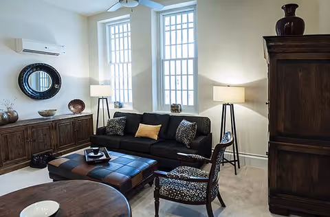 A cozy living room with a dark sofa adorned with patterned and solid pillows, a large ottoman coffee table, two patterned armchairs, two tall floor lamps, a wooden cabinet with decorative bowls, a round mirror on the wall, and two tall windows letting in natural light.