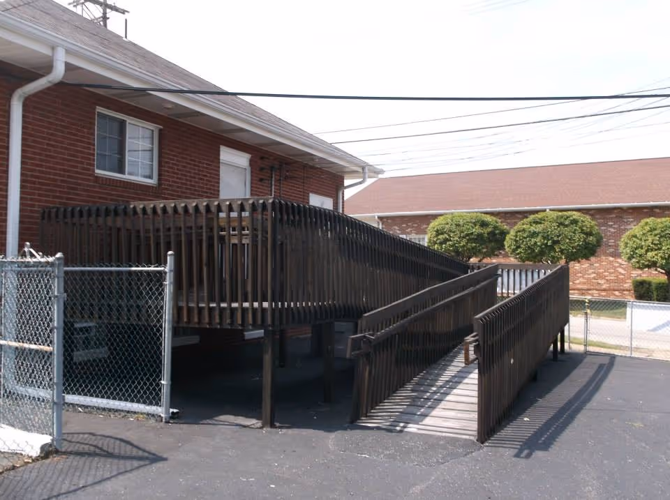 Wooden wheelchair ramp and deck leading to the entrance of a brick building, with a chain-link fence and trimmed shrubs nearby.