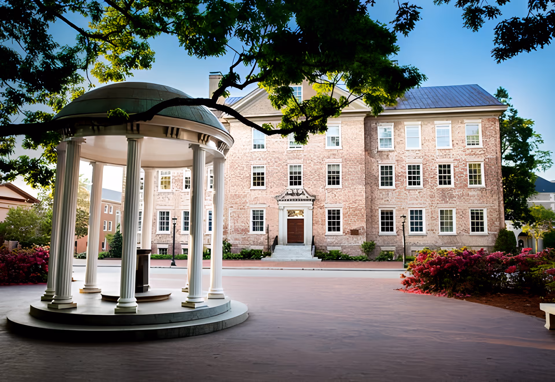 A historic brick building with multiple windows and a central entrance, viewed from across a paved area with a white gazebo featuring columns in the foreground. Trees with green leaves frame the top of the image, and there are bushes with red flowers on the right side.