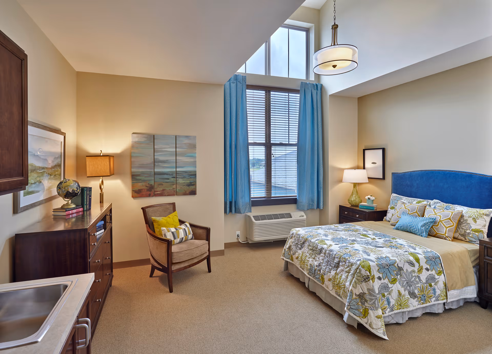 A cozy bedroom with a large window covered by blue curtains, a bed with floral bedding and multiple pillows, a nightstand with a lamp and decorative items, a wooden dresser with a globe and books, a comfortable armchair with cushions, and a small sink in the corner.