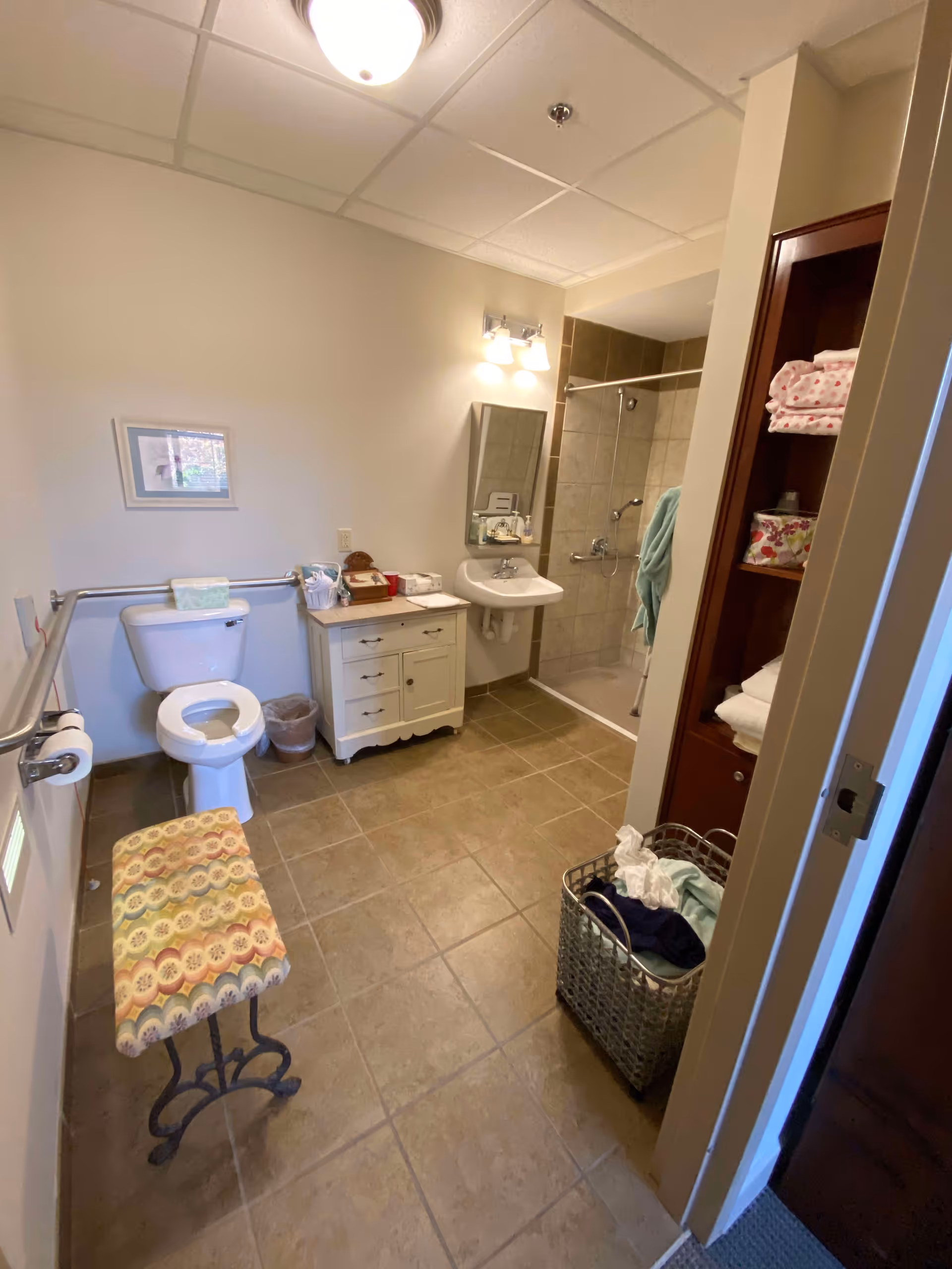 A spacious bathroom with a toilet, a small white cabinet with toiletries, a wall-mounted sink with a mirror and light above it, and a walk-in shower with a glass door. There is a patterned cushioned bench near the door, a laundry basket with clothes, and an open shelving unit with towels and other items. The floor is tiled and the walls are painted light beige.