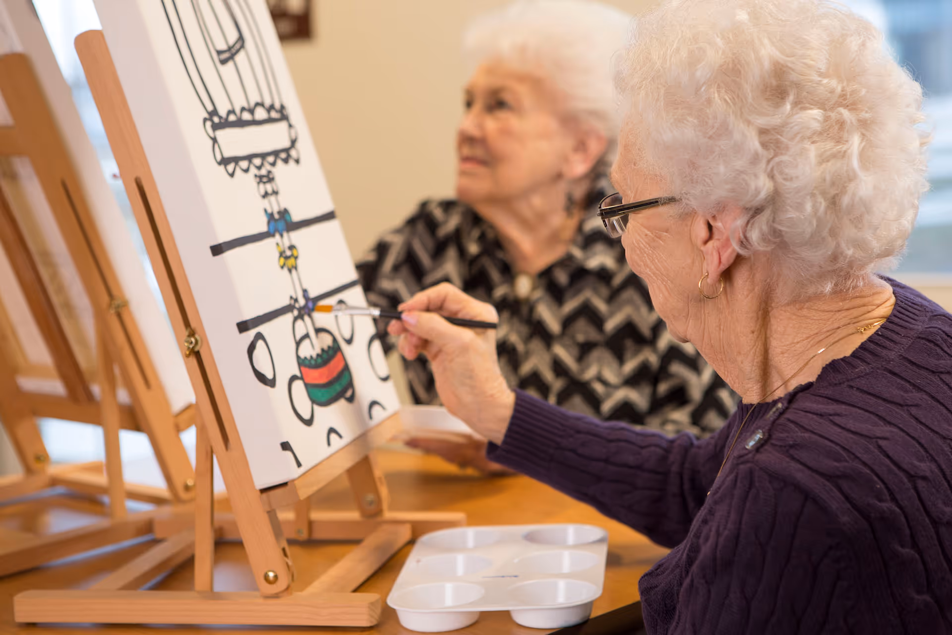 Two elderly women engaged in painting on canvases set on wooden easels. One woman is actively painting with a brush, while the other looks on. They are seated at a table with a paint palette in front of them.