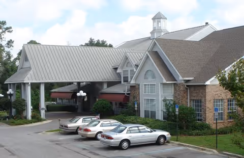 Exterior view of a senior living facility named The Waterford at Carpenter's Creek, showing a covered entrance with a metal roof, a brick building with large windows, several parked cars, and surrounding greenery.