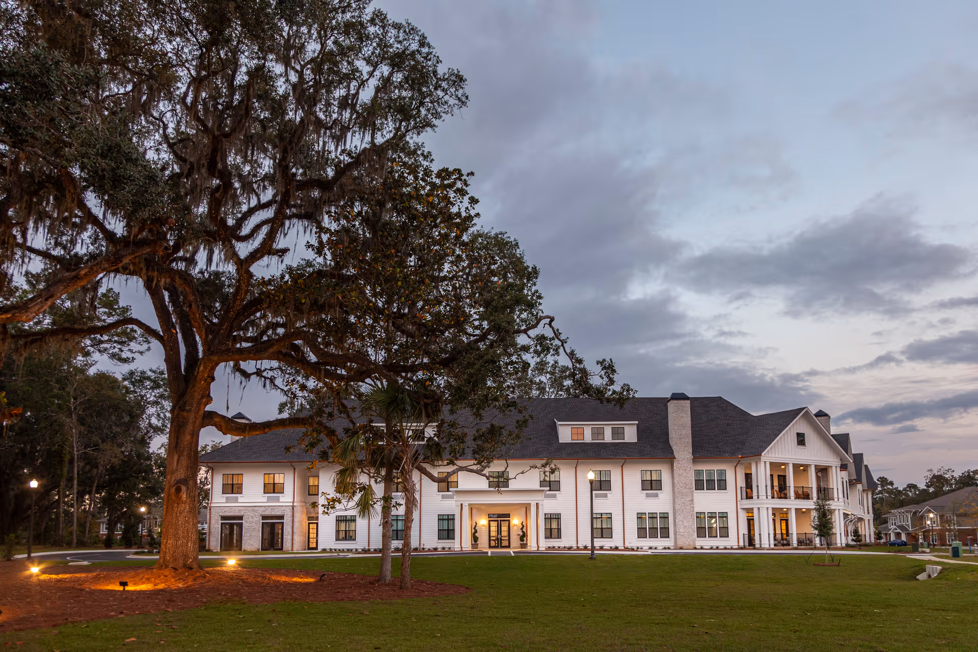 A large, two-story white building with multiple windows and a covered entrance, surrounded by a well-maintained lawn and large trees under a cloudy sky at dusk.