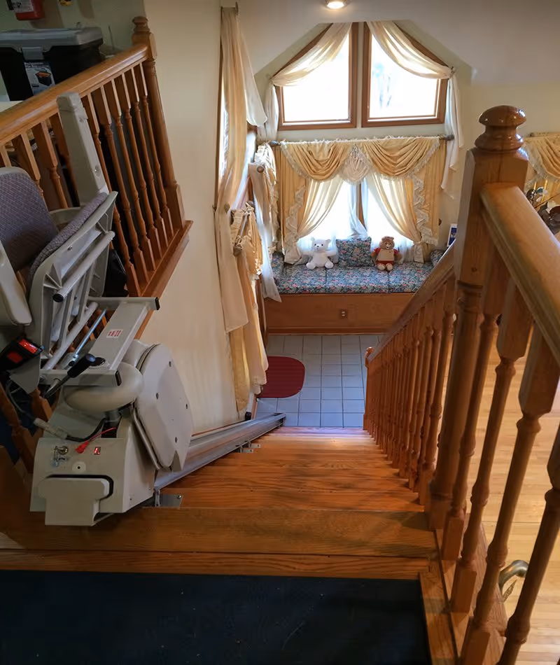 Wood staircase with a stairlift descends toward a sunlit window nook with stuffed animals and curtains.