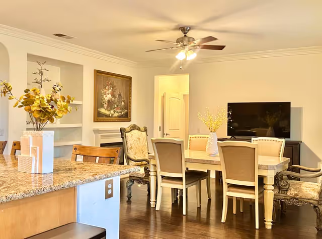 A bright and cozy interior space featuring a dining area with a rectangular table surrounded by six chairs, two of which have ornate armrests. A large flat-screen TV is mounted on a stand against the wall. To the left, a granite countertop with wooden chairs is visible, along with a vase holding yellow flowers. The room has wooden flooring, a ceiling fan with lights, and a framed floral painting on the wall above a white fireplace mantel.