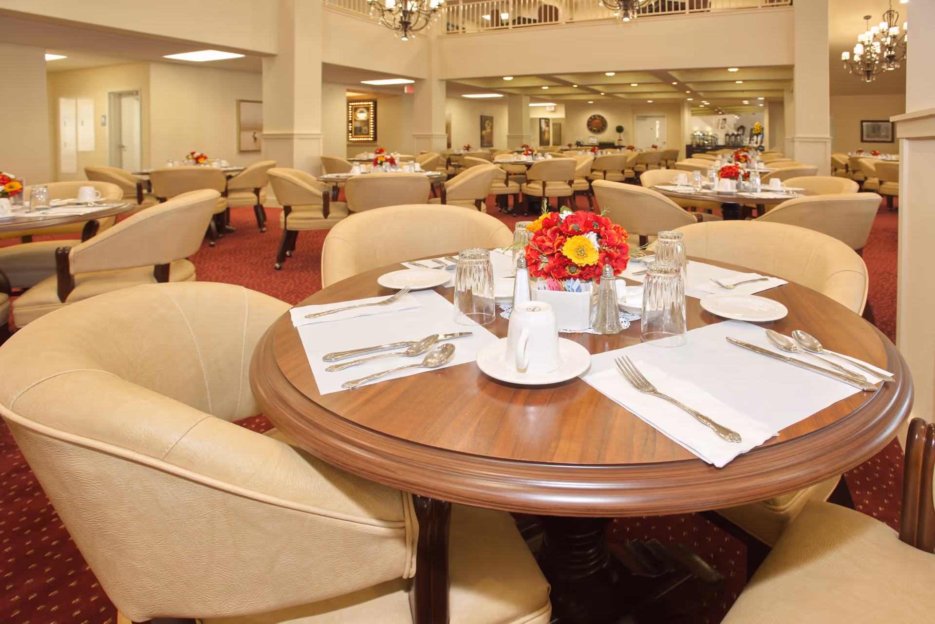A spacious dining room with round wooden tables set with white placemats, plates, silverware, glasses, and a centerpiece of red and yellow flowers. The room features beige upholstered chairs, red carpet flooring, chandeliers, and soft lighting, creating a warm and inviting atmosphere.