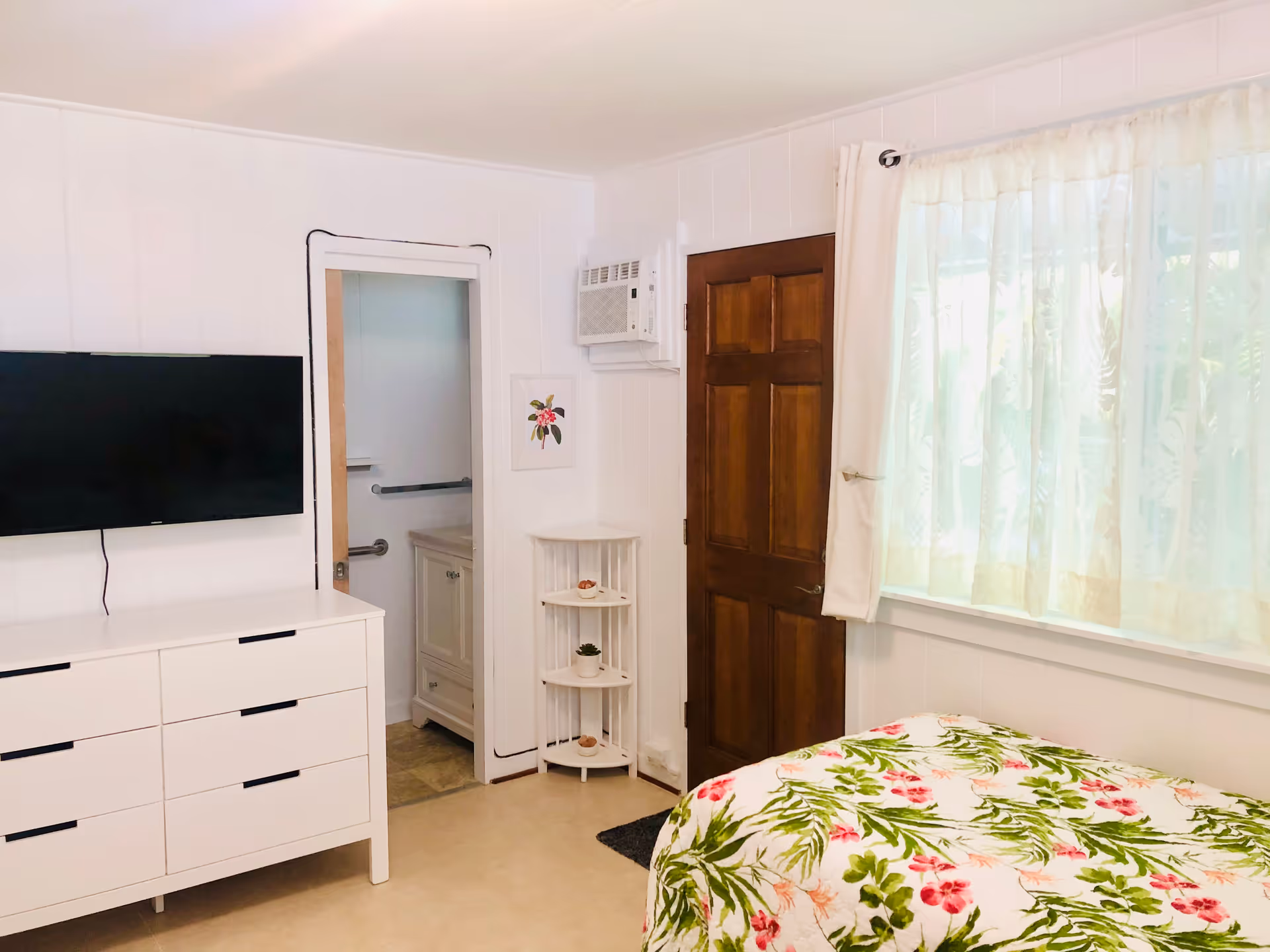 A bright bedroom with white walls and a large window covered by sheer curtains. There is a bed with a floral patterned bedspread featuring green leaves and pink flowers. A white dresser with multiple drawers is positioned against the wall, with a flat-screen TV mounted above it. A wooden door and a small corner shelf with decorative items are also visible. An open doorway leads to a bathroom with a vanity and grab bars.