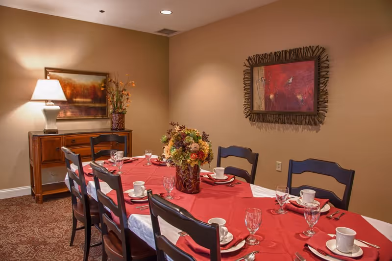 A dining room with a long table set for six people, featuring a red tablecloth, white plates, cups, glasses, and silverware. A floral centerpiece is in the middle of the table. The room has beige walls with two framed paintings and a wooden sideboard with a lamp and a vase of flowers.