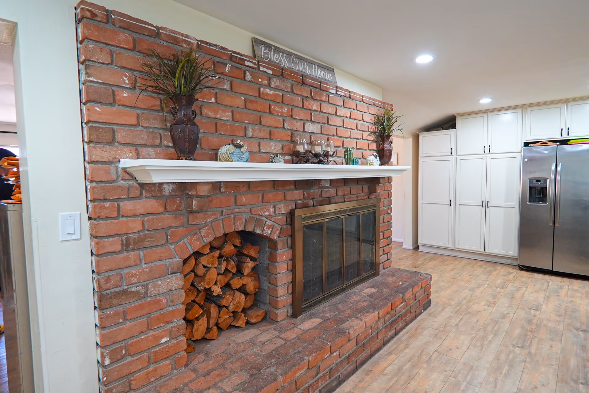 Interior view of a room featuring a large brick fireplace with a white mantel decorated with plants, candles, and decorative items. To the right of the fireplace, there are white cabinets and a stainless steel refrigerator. The floor is wooden, and the ceiling has recessed lighting.