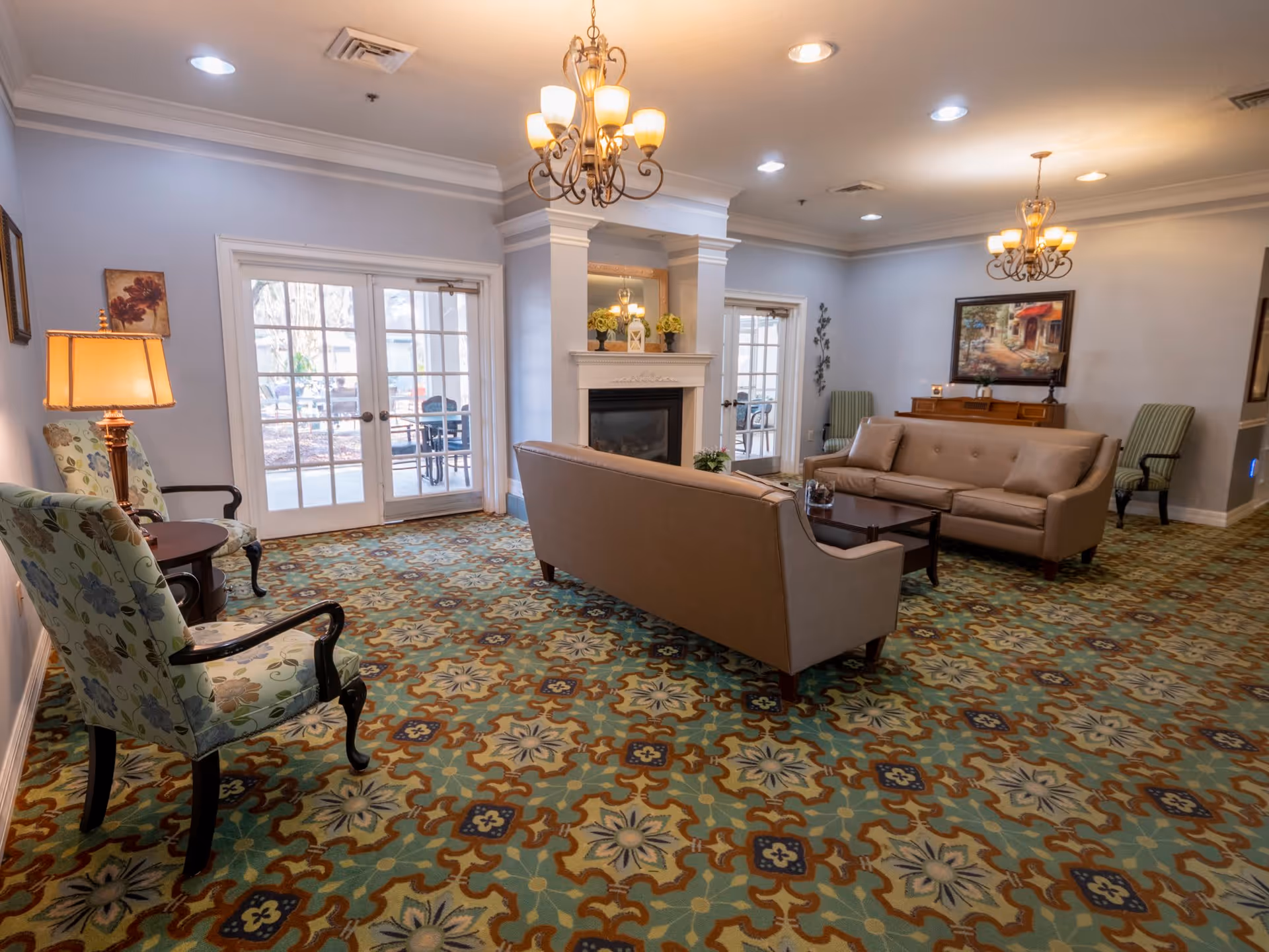 A cozy living room with patterned carpet, two beige sofas facing each other around a dark wooden coffee table, floral upholstered armchairs, a fireplace with decorative items on the mantel, and glass doors leading to an outdoor patio.