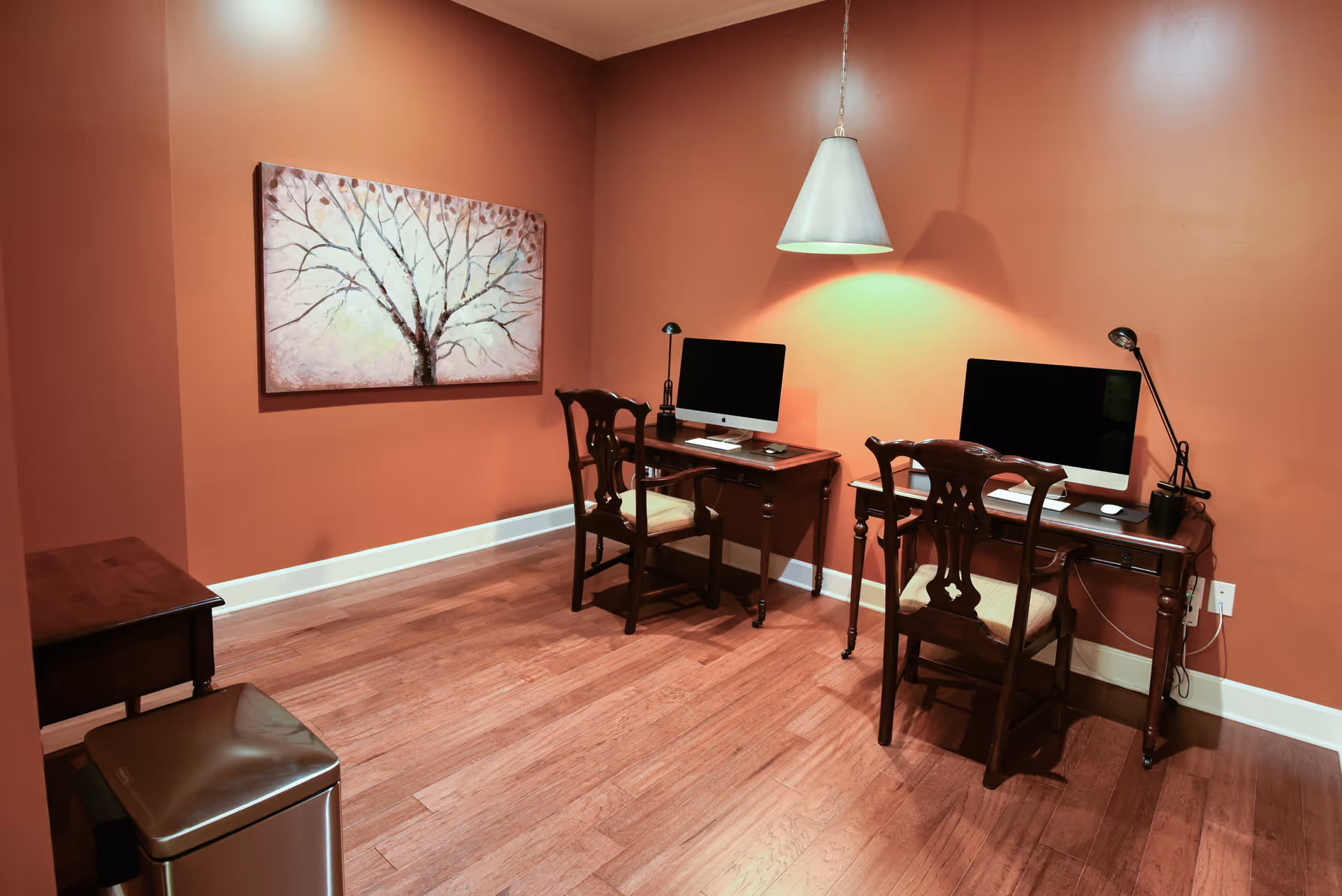 A small office or computer room with two wooden desks, each with a computer and a wooden chair. The walls are painted a warm orange color, and there is a painting of a tree on the left wall. A hanging light fixture illuminates the desks, and the floor is wooden.