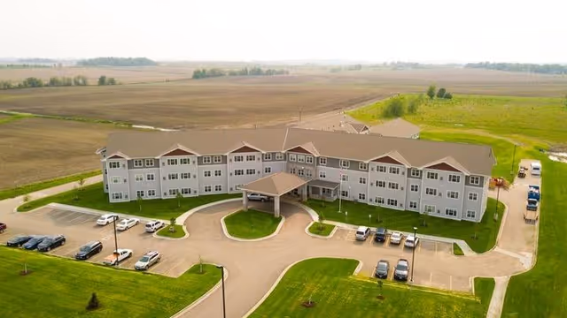 Aerial view of a large, three-story senior living facility named Stoney Point Meadows surrounded by green lawns and open fields. The building has a covered entrance with a circular driveway and multiple parked cars in the parking lot.