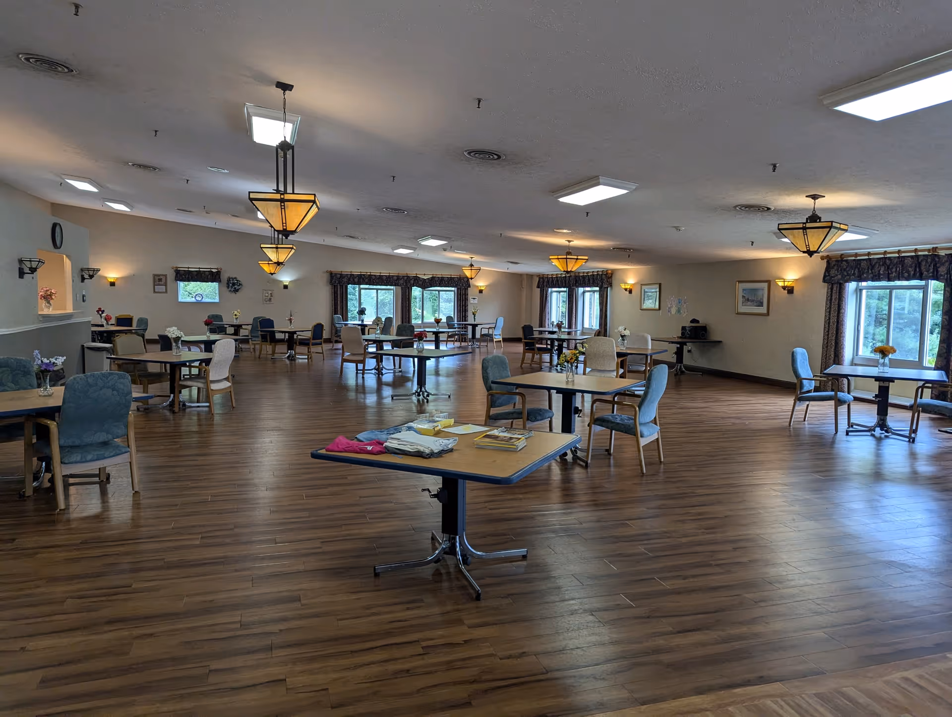 A spacious dining room with multiple tables and chairs arranged throughout the room. The floor is wooden, and the room is well-lit with ceiling lights and hanging pendant lamps. Windows with curtains allow natural light to enter, and there are small flower arrangements on some tables. The room appears clean and organized, suitable for communal dining or activities.